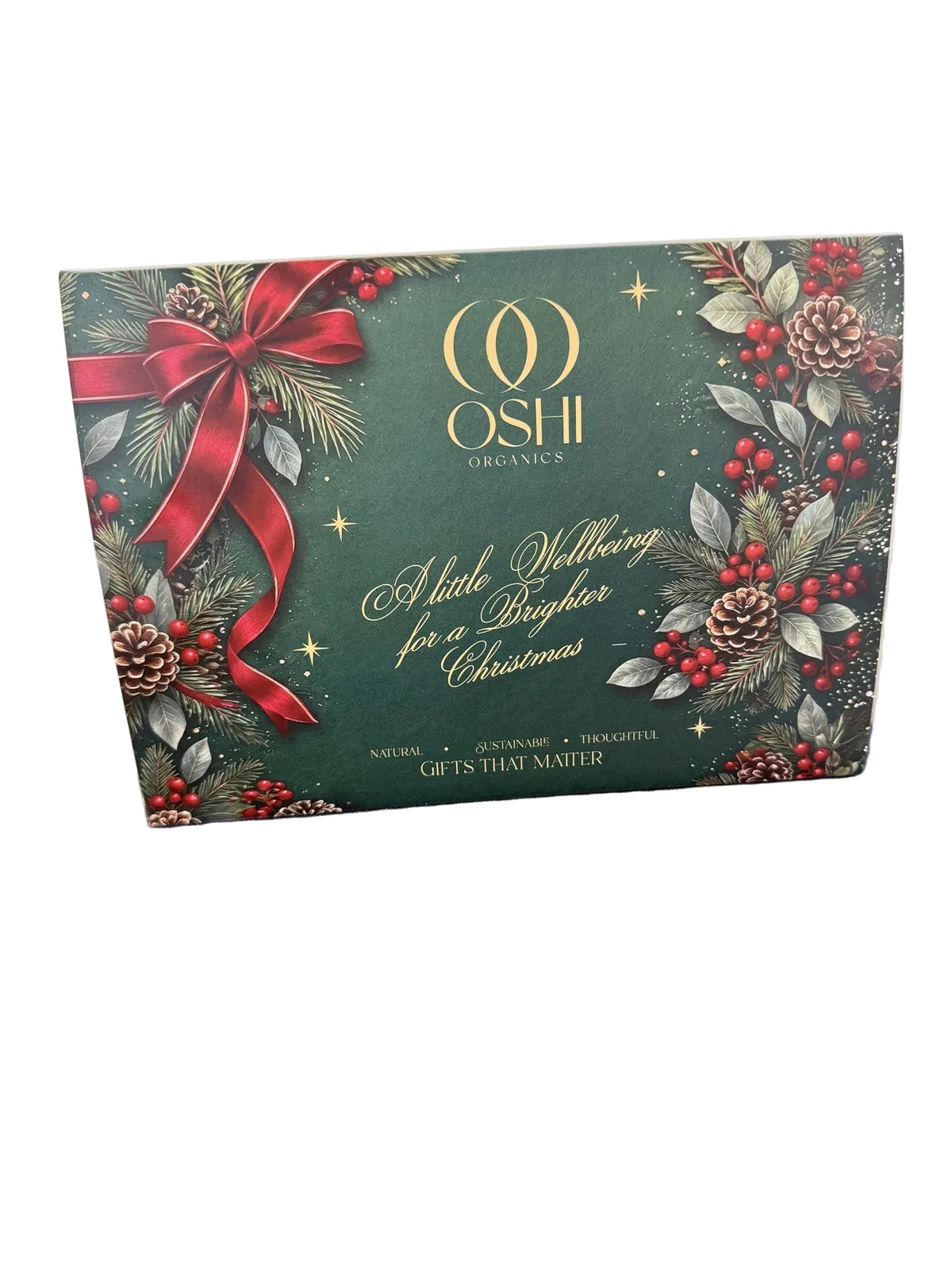 Oshi Organics 'A Little Nettleing for a Brighter Christmas' gift box, featuring a dark green holiday design with a large red bow, pinecones, and red berries, resting on a blue textured surface.